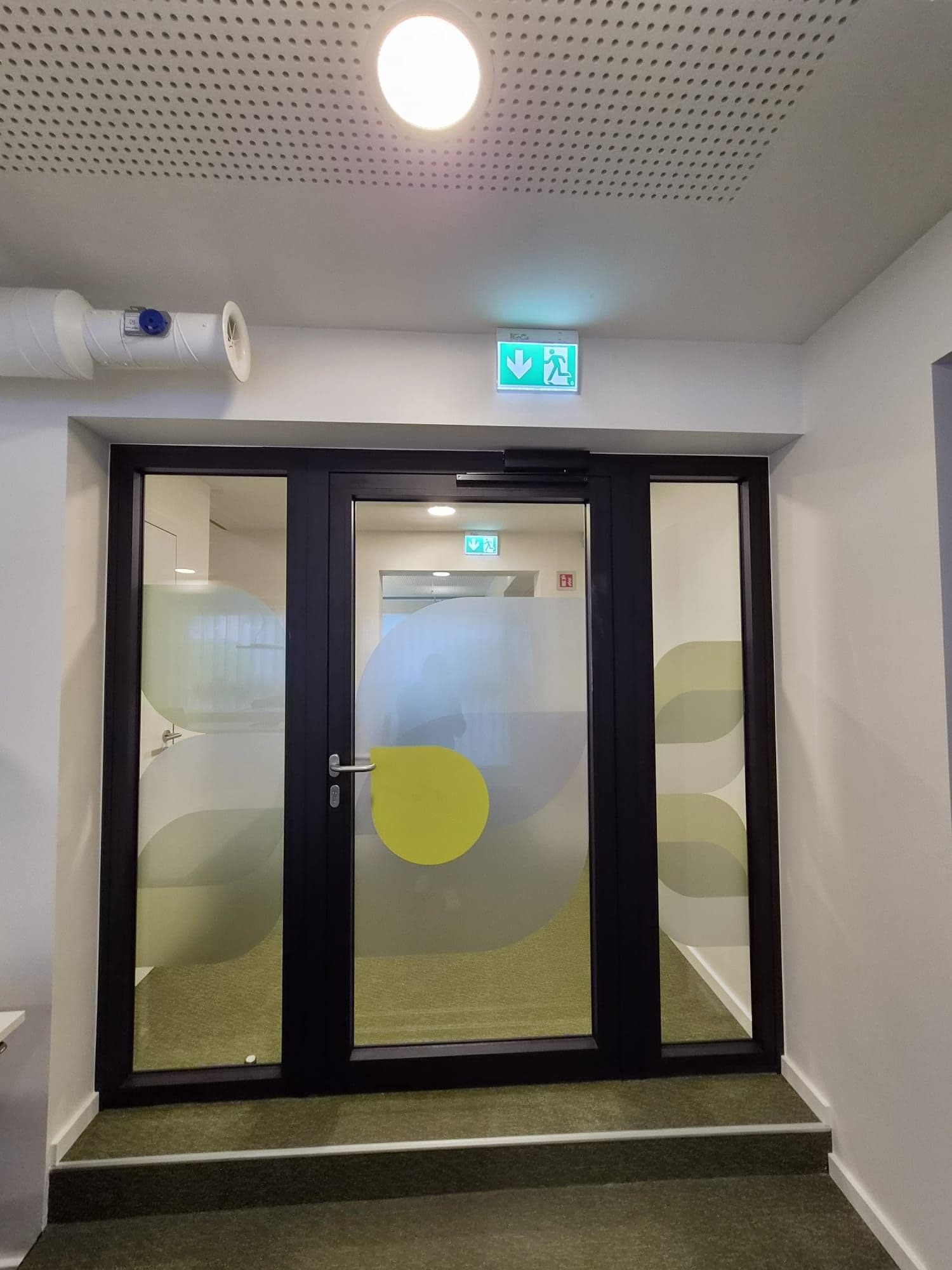 Black glass door with green patterns in a lit corridor, with emergency exit
